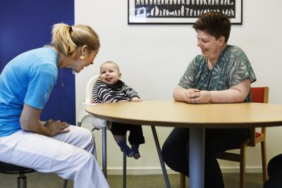 Billedet viser en dreng på 6 måneder og hans mor sidde ved et bord sammen med en AVT-terapeut til en AVT-session. Drengen stikker tungen ud og mor og AVT-terapeut griner.