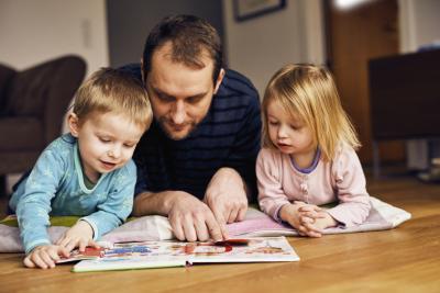 Far på gulvet med to børn. Drengen har en blå bluse på. Pigen har en lyserød bluse på. De kigger i en bog.